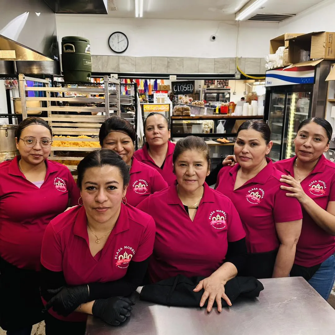 seven Hispanic women in Plaza Morelia shirts posing for a team photo in the kitchen of Plaza Morelia
